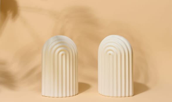A small, white ceramic decorative arch with vertical ribbed texture stands on a beige surface with blurred leaf shadows