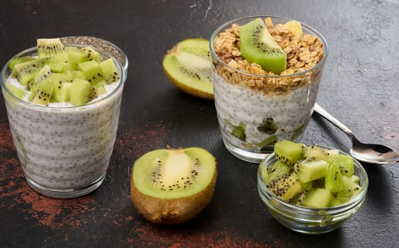 Two glasses of chia seed pudding, one topped with diced kiwi and the other with granola and banana chips, are on a dark surface with ingredients