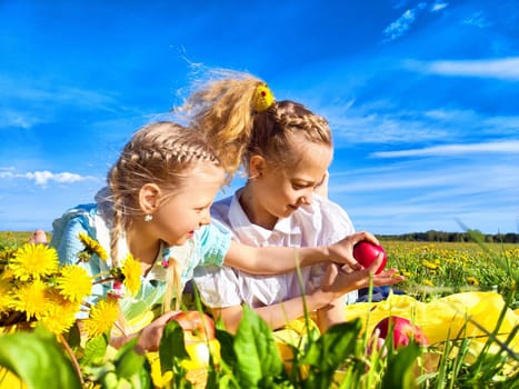 The girls are sitting in a lush green field adorned with bright flowers, happily painting colorful eggs on a sunny day.