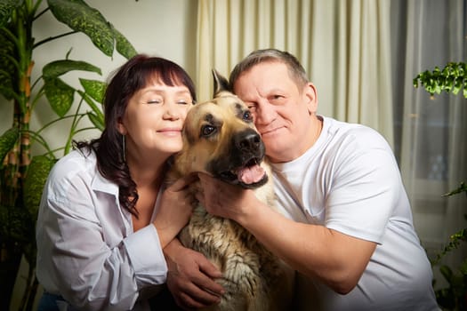 A middle-aged couple joyfully poses with their German shepherd inside their apartment, surrounded by indoor plants, showcasing their family bond.
