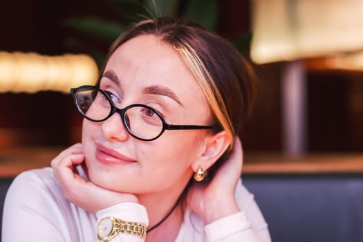 Portrait of a satisfied beautiful young woman wearing glasses in a restaurant looking aside with a smile while waiting for an order. High quality photo