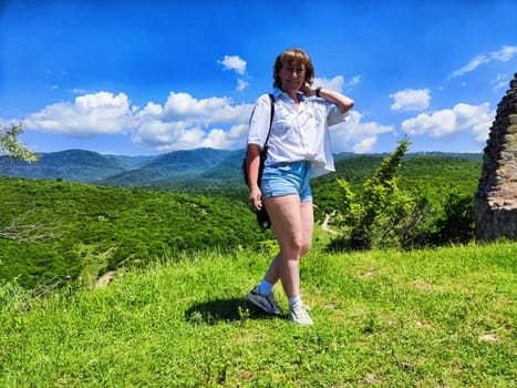 A female traveler stands on green grass with mountains in the background, enjoying a sunny day while exploring the outdoors.
