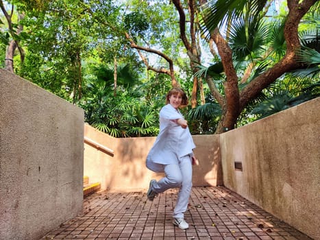 A mature woman with blonde hair strikes a playful pose while exploring a scenic outdoor area surrounded by tropical foliage.