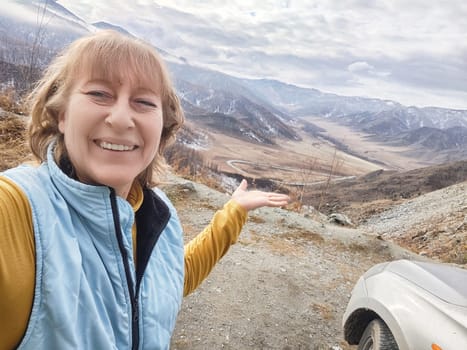 Blonde woman happily takes a selfie while pointing at a breathtaking valley view during her travel trip in the mountains.