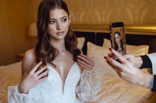 Bridesmaid taking photos of beautiful bride on her phone. Bride with makeup and hairdo in lace dress posing in front of phone camera. Wedding day morning.