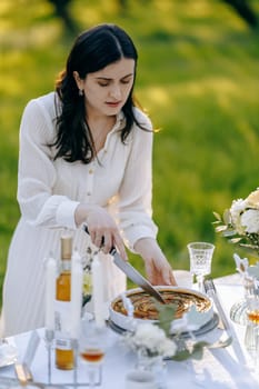 A woman is skillfully cutting a delicious pie on a beautiful table set up outside, enjoying the fresh air and sunshine