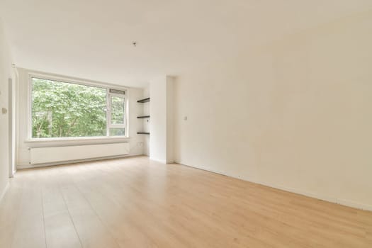An empty, minimalist interior room featuring large windows allowing natural light to fill the space. The wooden floor complements the bright atmosphere.