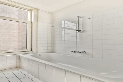 A bright and minimalist bathroom showcasing a bathtub, large window, and white tiled walls. The interior emphasizes simplicity and cleanliness.