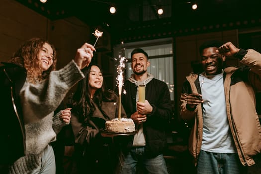 Group of friends joyfully celebrating together at night, holding a cake with sparklers. Laughter and excitement fill the air as they share this memorable moment.