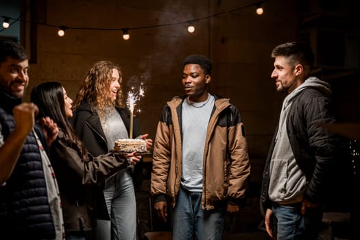 Group of friends enjoying a nighttime celebration outdoors. One person holds a decorated cake with sparklers while others cheer and smile in a cozy atmosphere.