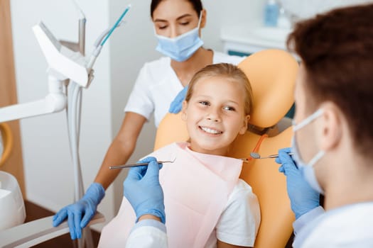 Modern dental examination and oral cavity treatment in children. Cheerful little kid in medical chair looks at millennial male doctor in white coat, protective mask and rubber gloves works with team