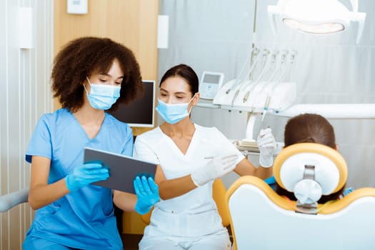 Modern technology and X-rays in dental office. Young european and african american female doctors in protective masks and rubber gloves, treat child in chair and look at tablet in clinic with tools