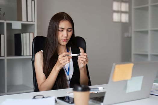 A businesswoman sits at her office desk, feeling unwell, checking a thermometer. The modern office setting includes a laptop and coffee cup.