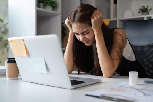 A young businesswoman feeling tired and stressed at her desk, surrounded by work materials in a contemporary office setting.