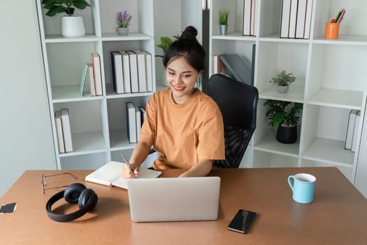 A young woman diligently doing her homework at home, using a laptop and surrounded by books in a modern study environment.