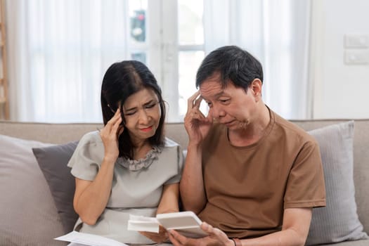 An elderly couple sitting on a couch, looking stressed while reviewing bills and financial documents at home.