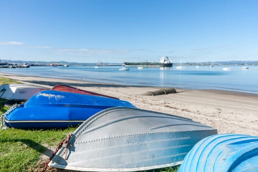 Group of upturned dinghies pulled up on grass edge to beach with boats and port activity in background to Pilot Bay Mount Maunganui.