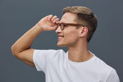 Smiling young caucasian man with brown hair and glasses wear white t-shirt looking right side on plain grey background. Positive emotion, casual style, mental health and digital detox concept.