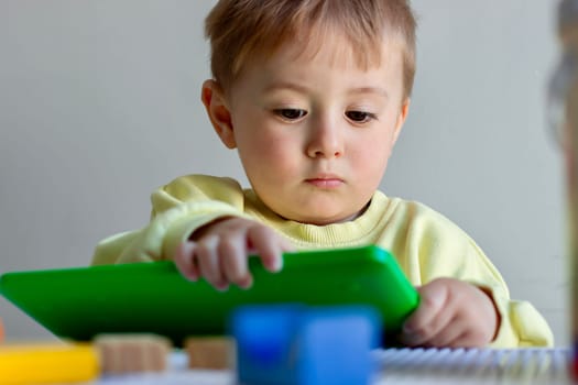 Little serious two-year-old boy in a yellow sweater playing with a green tablet at a children's table. High quality photo