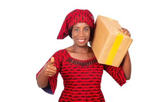 beautiful african adult woman wearing red loincloth standing holding a cardboard box on her shoulder and making a positive gesture with hand thumbs up smiling and happy for success