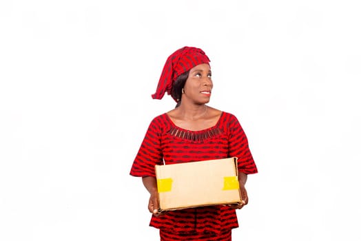 happy beautiful mature african woman standing and dressed in red loincloth suit looking aside while holding a cardboard box on white background