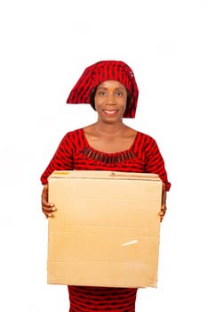 happy beautiful mature african woman standing and dressed in red loincloth suit holding a big cardboard box on white background