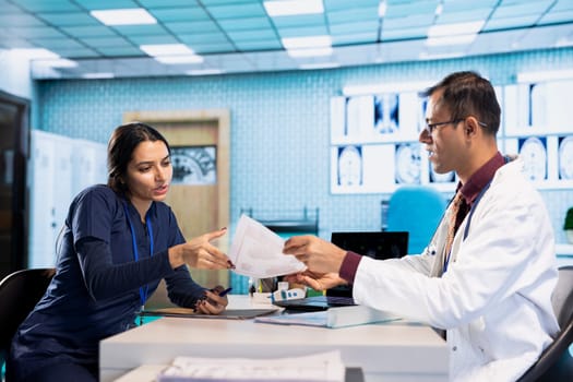 Indian expert explaining diagnostics through test results with a male patient at medical office, doing scan interpretation at check up with young man. Healthcare services on insurance.