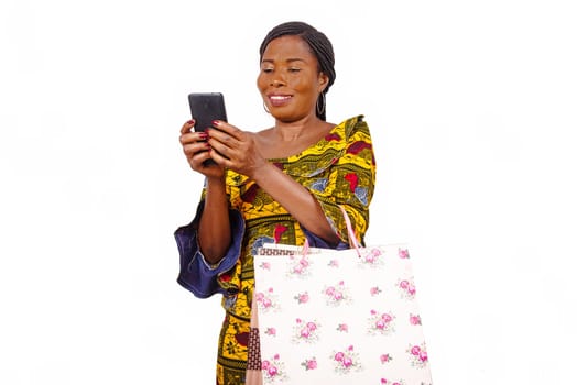 Portrait of beautiful african adult woman holding colorful paper bags looking at a message or videos on her mobile phone isolated on white background