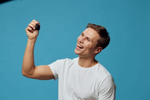 Happy young caucasian man wearing white t-shirt smiling and holding car keys showing excitement and freedom while isolated on blue background. Digital nomad concept with positive emotion and casual style.