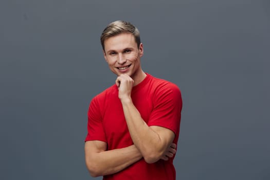 Happy young man with short neatly styled hair and light skin, wearing a red casual t-shirt, poses confidently against plain gray background, smiling warmly and looking directly at the camera, casual style, studio shot, positive mood