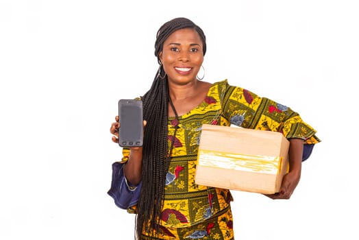 beautiful african woman standing in traditional outfit on white background showing cellphone smiling and carrying a cardboard.