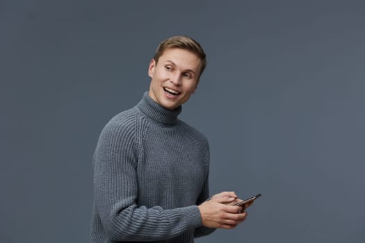 Happy man with casual gray sweater holding mobile phone, smiling and looking at camera, neutral gray backdrop, youthful style, positive mood, indoors, people lifestyle, modern fashion.