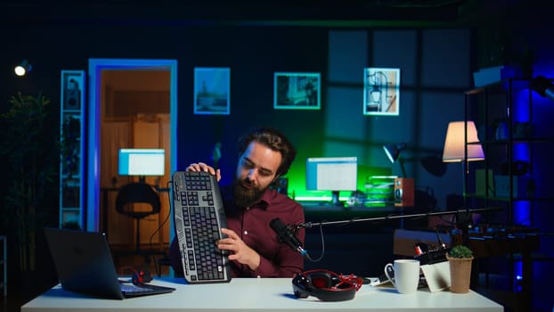 Man in studio reviewing gaming keyboard received from sponsoring brand. Online show host paid to promote bluetooth wireless computer peripheral to viewers during live broadcast, camera B