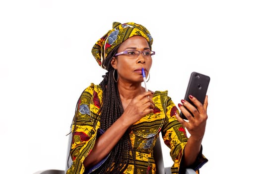 Beautiful adult woman in colorful loincloth is sitting holding a pen in her mouth, thinking while looking at the cell phone.