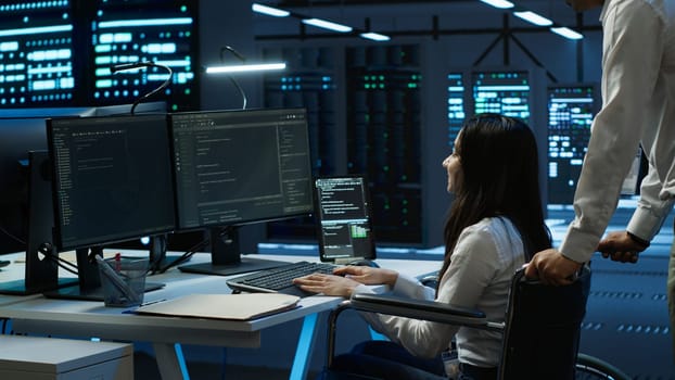 Person with paraplegia helped by coworker in data center to arrive at desk, coding on PC. Woman in wheelchair doing maintenance on server room infrastructure rackmounts, camera A