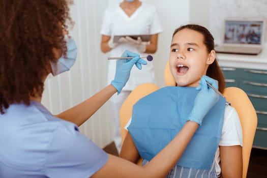 Medical treatment to small patient at modern dental clinic. Young african american dentist in uniform, protective mask and rubber gloves treats teeth of latin girl with open mouth in chair, cropped