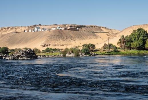 Granite rocks in the fast flowing water of the River Nile near Aswan Egypt with resort on hilltop