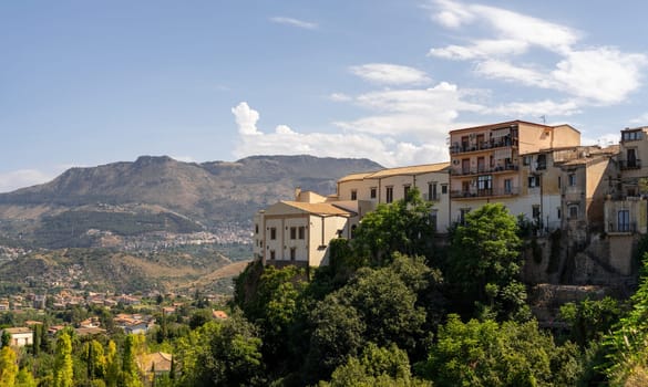 Old buildings perched on hill, surrounded by lush greenery, overlooking vast scenic valley with distant mountains near Palermo city, Monreale town, Sicily, Southern Italy