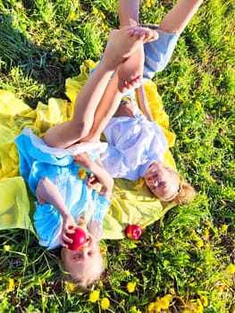 Two girls lay on a bright yellow blanket in a grassy field, playfully holding apples and enjoying a sunny summer day outdoors.