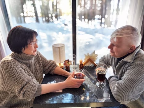 A couple shares a moment at a table, sipping drinks and engaging in conversation. The warm light contrasts with the snowy landscape outside, creating a cozy atmosphere.