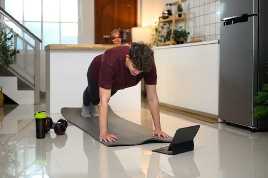 Active man doing plank exercise on mat and following an online workout on digital tablet.