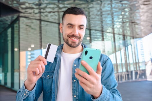 Happy young man using his smartphone while holding a credit card. He smiles with excitement, standing outside a glass building, enjoying an online shopping or payment experience.