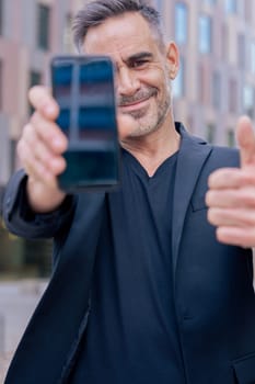 Smiling man holding smartphone towards the camera and giving a thumbs up outdoors. He wears a black jacket and stands in a modern urban space. Concept of approval, technology, and confidence. Vertical