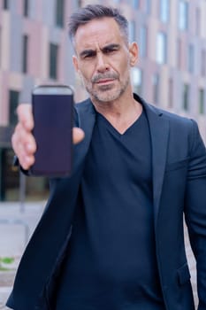 Confident Caucasian man in black outfit holding smartphone toward the camera with serious face. Business, technology, and communication concept in an urban environment with modern architecture.