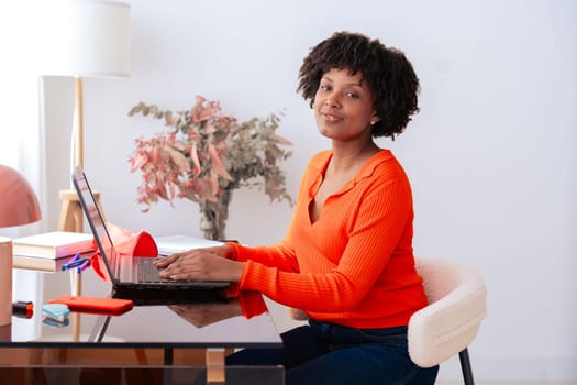 African American woman sitting at home office desk, working on laptop and looking at camera. Modern workspace with books, stationery, and decor. Remote work, productivity, technology, and confidence.