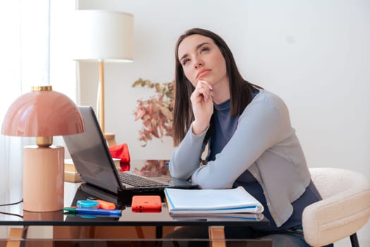 Caucasian woman sitting at desk, thinking while looking up. Laptop, notebook, and stationery on desk in modern home office. Creativity, concentration, decision-making, productivity, and contemplation.