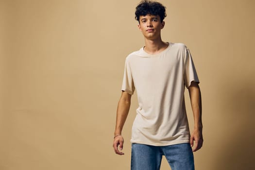 A stylish young man poses in a beige t shirt and blue jeans on a neutral brown backdrop, embodying a modern casual look suitable for lifestyle and fashion photography