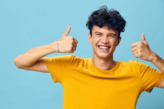 cheerful young man with curly hair giving thumbs up with both hands, wearing a vibrant yellow t shirt, set against a light blue background, radiating happiness and positivity