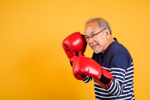 Portrait Asian old man wearing glasses wear two red boxing gloves studio shot isolated yellow background, smiling happy elderly man gray haired healthy fighter lifestyle concept