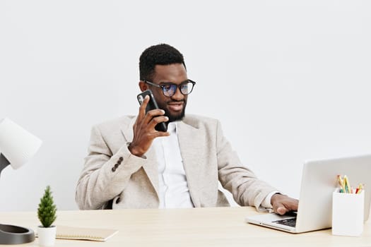 Smiling young African American man in smart casual clothing working at a contemporary office desk, communicating via mobile phone, using a laptop, surrounded by minimal decor, natural light, indoor workspace, conveying business success and professionalism.